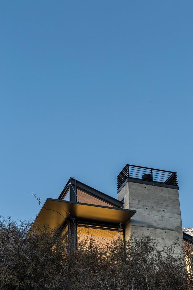 Upward-looking exterior view of a contemporary multi-level house at dusk, featuring a prominent raw concrete tower with a rooftop terrace enclosed by dark horizontal metal railings. Adjacent pitched roof forms and warm timber-clad wall panels sit beneath a broad projecting canopy, with dense shrubs in the foreground and a clear blue evening sky.