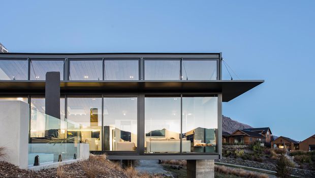 Exterior view of a contemporary, elevated glass-fronted house with a strong horizontal flat roof and projecting cantilevered canopy. Floor-to-ceiling glazing reveals a bright open-plan interior, while the structure sits above a landscaped sloping site with mountain terrain and neighboring houses visible in the background.