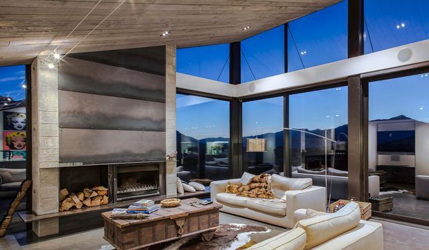 A spacious contemporary living room with floor-to-ceiling corner glazing overlooking a mountainous urban setting. The room features a large white sectional seating arrangement, a substantial exposed fireplace with stacked firewood, a timber coffee table, and a warm wood-clad ceiling. The high glazing and evening blue light create an airy yet refined atmosphere.