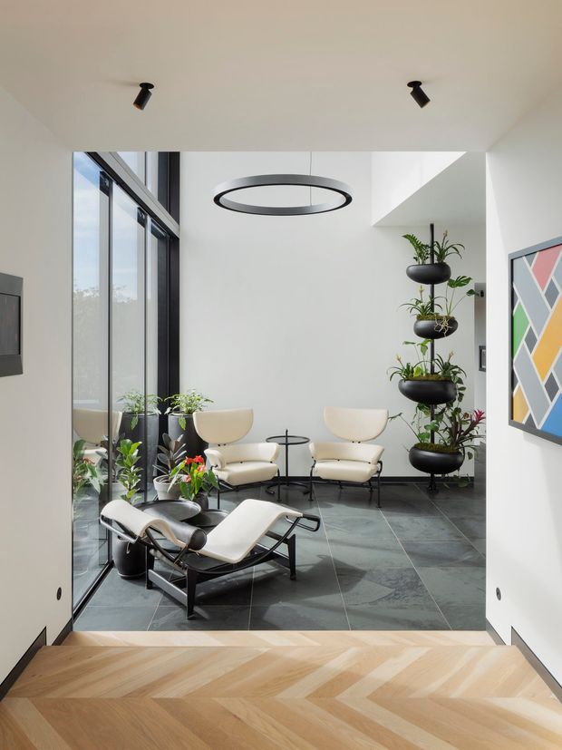 A refined modern sitting area viewed from a light timber herringbone-floored threshold. The room has large black-framed floor-to-ceiling glazing, dark grey tiled flooring, white walls, two cream lounge chairs, a matching chaise lounge, small black side tables, potted plants, a circular pendant light, and colourful geometric wall art.