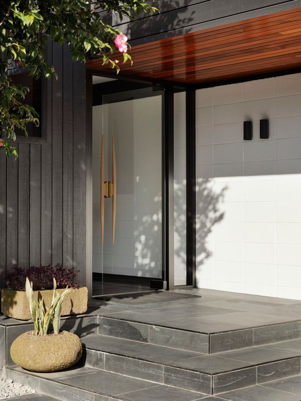 A contemporary residential entry facade viewed from outside, featuring a black vertical timber-clad wall, a glass entry door with dark frame, a white tiled garage or entry wall, and a warm timber-lined soffit. Broad dark stone steps and paving lead to the doorway, with sculptural planters and leafy landscaping framing the entrance.