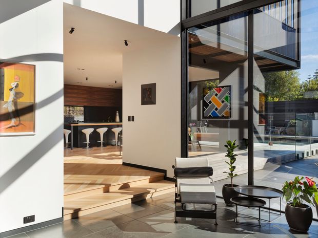 A bright, double-height modern interior centred on an open living and entry area. Full-height black-framed glazing overlooks a pool terrace, while broad timber steps lead up to a kitchen or bar zone with dark timber cabinetry and sculptural white stools. The space includes a leather lounge chair, small round tables, indoor plants, contemporary artwork, polished stone-like floor tiles, and strong geometric sunlight and shadow.