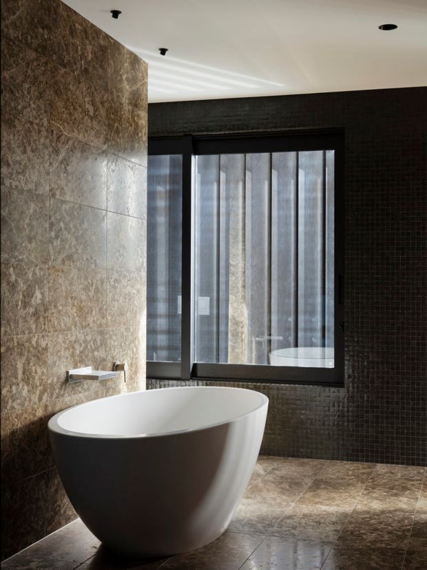 A sophisticated contemporary bathroom featuring a freestanding oval bathtub, a wall-mounted bath filler, large mottled stone-look wall tiles, dark small-format tiled walls, and matching stone-look floor tiles. Black-framed windows with sheer coverings provide soft natural light, while recessed ceiling lights add subtle illumination.