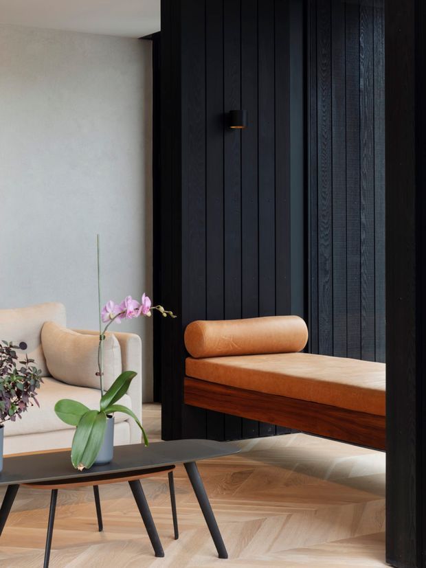 A refined contemporary interior corner featuring a dramatic black vertical timber wall and a cantilevered built-in bench with a warm tan leather seat and cylindrical bolster. A cream upholstered armchair, dark coffee table, potted orchid, and herringbone timber flooring complete the composition. The space is warm, high-contrast, and carefully detailed, with strong built-in joinery and sculptural furniture.