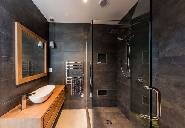 Contemporary bathroom with dark large-format wall tiles, a timber vanity with vessel basin, wall-mounted mirror in a timber frame, heated towel rail, and a frameless glass walk-in shower with dual shower fittings. The room has a refined, spa-like feel with warm timber contrasting against charcoal surfaces.