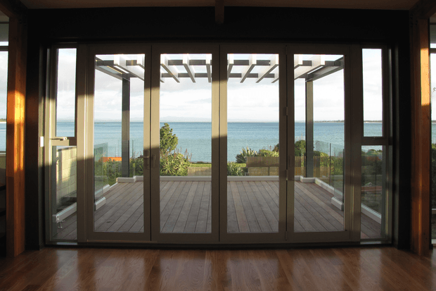 Interior view through large dark-framed glazed folding or sliding doors toward a timber deck, pergola structure, landscaped garden, and ocean beyond. The room has warm polished timber flooring and exposed dark timber framing, with a strong indoor-outdoor connection and coastal outlook.
