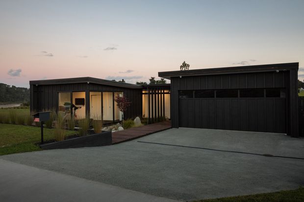 Exterior view of a low-profile contemporary home at dusk, featuring dark vertical cladding, a flat or very shallow roofline, a large black double garage, glazed living areas, a timber walkway, and landscaped planting beds.