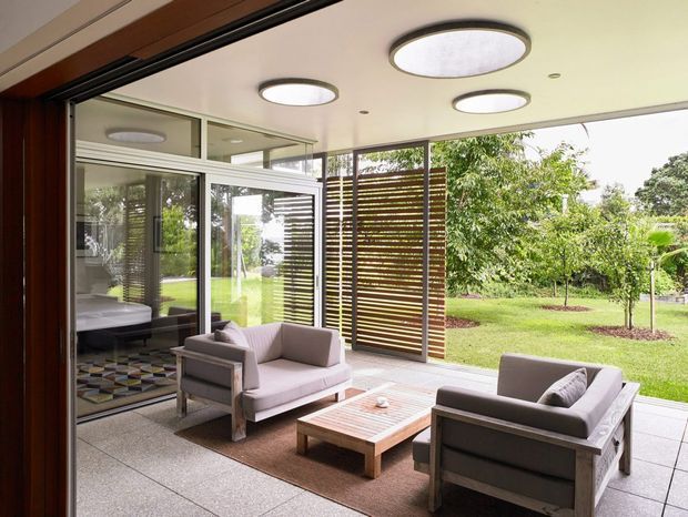 Bright contemporary indoor-outdoor living area with a seating arrangement of upholstered armchairs around a low timber coffee table. The space features large floor-to-ceiling glazed openings, timber-framed detailing, a pale smooth ceiling with circular recessed or flush-mounted lights, tiled flooring, and horizontal timber privacy screens overlooking a landscaped garden. A bedroom or adjacent room is visible through the glazing and reflection.