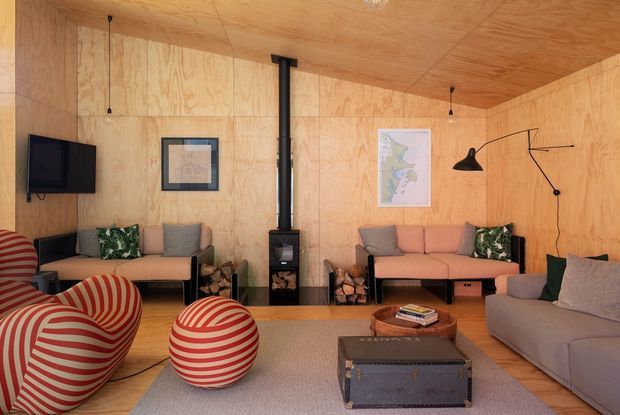 A warm, compact living room lined throughout with natural plywood, including the walls and ceiling. Two sofas and sculptural red-and-cream striped lounge chairs surround a low wooden coffee table and a large grey rug. A central black wood-burning stove with exposed flue is the room's focal point, complemented by wall-mounted lighting, framed artwork and a television.