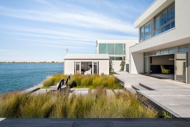 Exterior view of a contemporary waterfront residence with white rendered volumes, extensive glazing, broad timber decking, and an open garage or covered outdoor bay. The landscaped waterfront edge features ornamental grasses, a small pavilion, seating, and direct access toward the water.