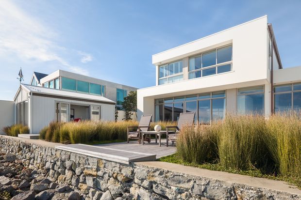 Contemporary multi-level waterfront residence viewed from the rocky shoreline, with clean white rendered volumes, extensive floor-to-ceiling glazing, flat rooflines, and a timber deck bordered by ornamental grasses. A smaller adjoining structure and landscaped retaining wall are also visible.