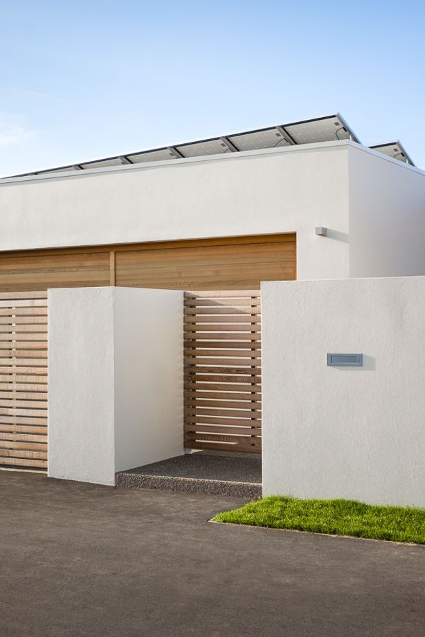 Street-level exterior view of a clean-lined contemporary residence, featuring white rendered walls, a flat roof with visible rooftop solar panels, a timber garage door, and a partially screened pedestrian entry with horizontal timber slats. Dark asphalt paving and a narrow strip of lawn sit in front of the facade.