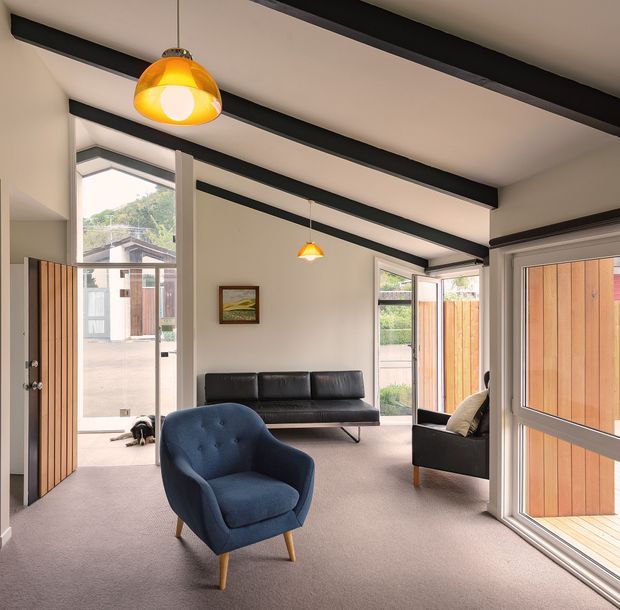 Bright, airy living room with high ceilings, exposed dark ceiling beams, warm pendant lights, carpeted flooring, and extensive glazing. The room contains a blue upholstered armchair, black leather sofa, and additional seating, with timber fencing and a residential exterior visible through the windows and open doors.