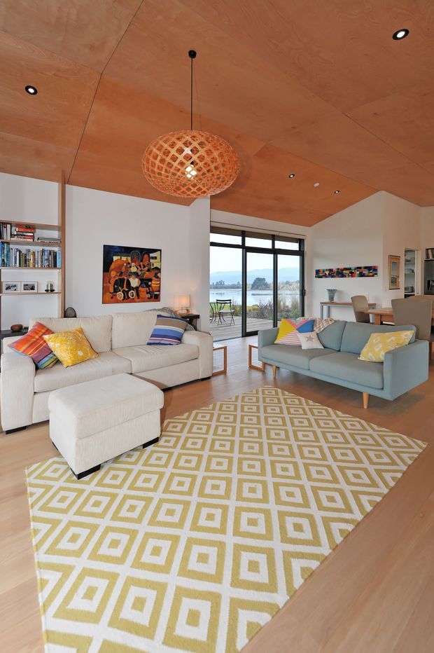 A bright, inviting living room with a warm plywood-lined ceiling, pale timber flooring, white built-in shelving, and large black-framed sliding glass doors opening to a water view. Two sofas, a matching ottoman, colourful cushions, a geometric area rug, artwork, and a woven pendant light create a relaxed contemporary setting.