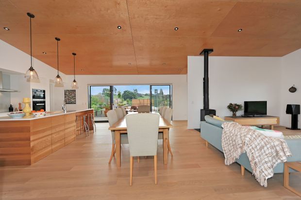 Open-plan contemporary living, dining and kitchen area with warm timber flooring and a timber-lined ceiling. The kitchen features extensive timber cabinetry, a pale benchtop, integrated appliances and pendant lighting. A timber dining table separates the kitchen from the lounge, which includes a sofa, television and freestanding black wood heater. Large glazed doors open to an outdoor area, creating a bright, connected interior.