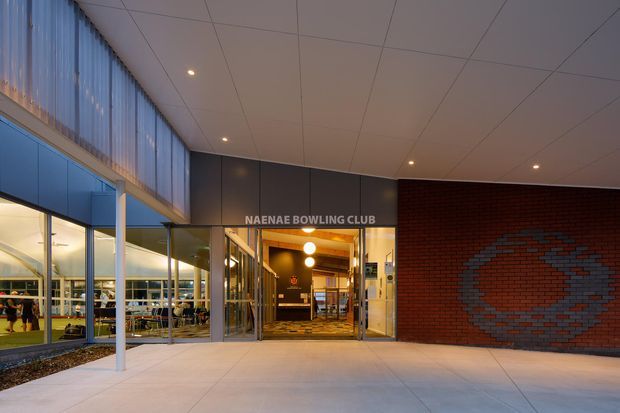 Exterior entrance view of the Naenae Bowling Club, showing a modern civic/commercial building with glazed walls, a covered entry canopy, illuminated reception foyer, and a red brick feature wall with a blue geometric pattern.