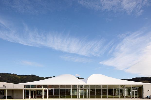 Wide exterior view of a low, modern building set against dark rolling hills. The building has extensive floor-to-ceiling glazed frontage with slender light-coloured frames and a distinctive sculptural white roof composed of smooth, flowing vaulted or domed forms. The scene is photographed in bright daylight beneath a blue sky.