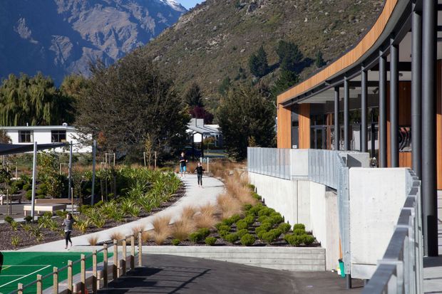 Exterior view of a contemporary building in a landscaped alpine setting. The building features a long curved roofline, extensive dark-framed glazing, warm timber cladding, exposed structural columns, and concrete retaining walls. Paved walkways, ornamental planting, and a green outdoor recreation surface occupy the foreground, with steep mountain terrain visible behind.