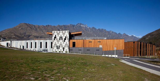 Wide exterior view of a contemporary low-rise building complex set against rugged alpine mountains. The building combines long white rendered volumes, a distinctive patterned vertical facade element, and warm timber-toned cladding, with paved access roads and open grass in the foreground.