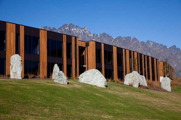 A long, low contemporary building with a dark glazed facade and repeated vertical timber-clad bays, set on a gently sloping landscaped lawn. Large pale sculptural rocks line the foreground, with rugged mountains clearly visible behind the building under a blue sky.