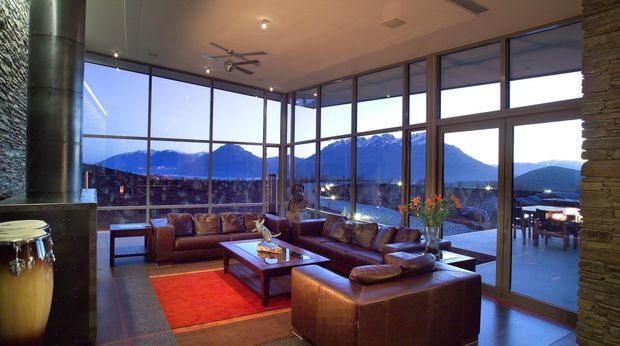 A spacious contemporary living room with extensive floor-to-ceiling glazing overlooking a dramatic mountain landscape. The room features dark leather sofas and armchairs arranged around a large timber coffee table, a vivid red area rug, a tall stone-clad fireplace wall, exposed cylindrical metal flue, recessed ceiling lights, and a ceiling fan. An outdoor terrace and dining area are visible beyond the glass doors.
