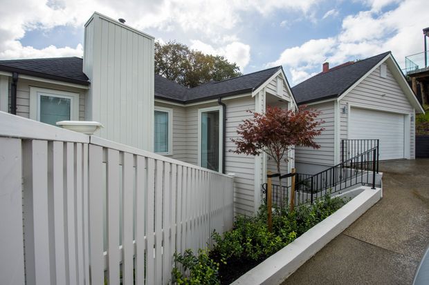 Exterior view of a compact residential property with multiple gabled roof forms, pale vertical and horizontal cladding, dark roofing, a raised white fence, and a gently sloped access path with black metal handrails. A small landscaped garden and Japanese maple sit beside the entry.