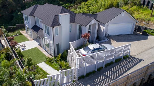 Aerial view of a multi-level suburban residence on a sloped site, with a grey weatherboard-clad house, multiple pitched roof sections, attached double garage, paved driveway, landscaped garden, and a white slatted boundary fence enclosing a parked car.