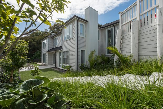 A modern two-storey residential home viewed from a landscaped garden. The exterior features light grey horizontal weatherboard cladding, white-framed windows, projecting upper-level volumes, and a tall narrow vertical element. Lush ornamental grasses, palms, broad-leaf plants, and a paved path surround the house, with a small outdoor terrace visible at the side.
