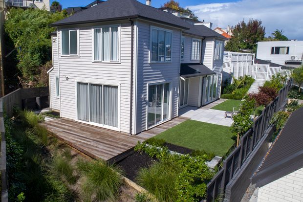 A two-storey suburban house with light weatherboard cladding, dark hipped roof, white-framed windows and doors, and a landscaped backyard. The property includes a timber deck, paved outdoor area, artificial lawn, garden beds, and white picket-style fencing.