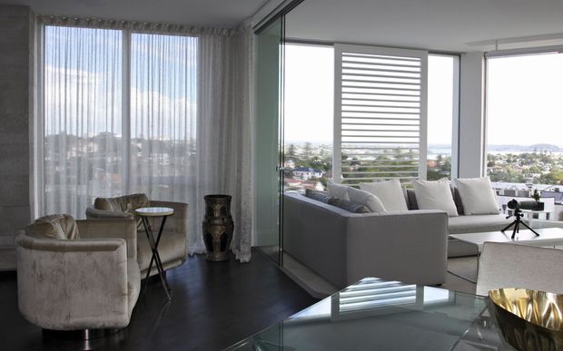 Bright contemporary living room with floor-to-ceiling glazing and sheer curtains, furnished with a large pale sectional sofa, plush armchairs, a small round side table, and a glass coffee table. The room opens toward a covered balcony or terrace with expansive city and water views, separated by a glass partition. Dark timber flooring, white walls, and horizontal exterior screening create a refined, airy interior.