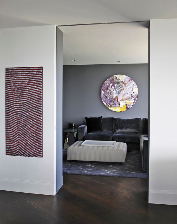 View through a wide doorway into a refined living room. The space features a dark charcoal velvet sofa, a large pale upholstered ottoman, a dark geometric rug, dark timber flooring, and a charcoal feature wall with a circular abstract artwork. White walls, concealed recessed lighting, and crisp architectural trim create a high-contrast, contemporary interior.