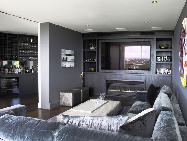 A sophisticated dark-toned living room with a large sectional sofa, ottoman, coffee table, built-in media wall, and linear fireplace. A fully fitted home bar with wine storage and glassware is visible to the left. The room features charcoal joinery and walls, plush grey upholstery, dark timber flooring, recessed lighting, and a view through the media wall glazing.