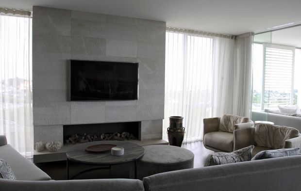 Bright contemporary living room with a large grey stone-clad fireplace wall, built-in linear firebox, wall-mounted television, low round coffee table, upholstered ottoman, and neutral sofas and armchairs. Floor-to-ceiling sheer curtains and extensive glazing create an airy, refined setting.