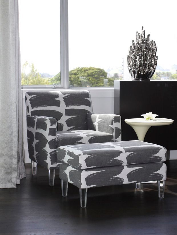 Bright contemporary seating corner with a patterned upholstered armchair and matching ottoman, positioned beside large windows. A black storage cabinet, sculptural metallic decor, sheer curtains, and a small white side table complete the monochrome interior.
