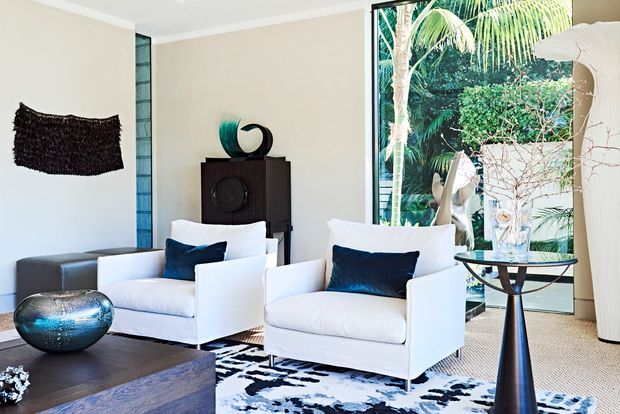 A bright, refined living room with two white upholstered armchairs, dark blue cushions, a dark timber coffee table, sculptural side table, patterned rug, and dark wood cabinetry. Full-height glazing opens the room visually to lush tropical foliage outside, while neutral walls and a textured dark wall artwork create contrast.