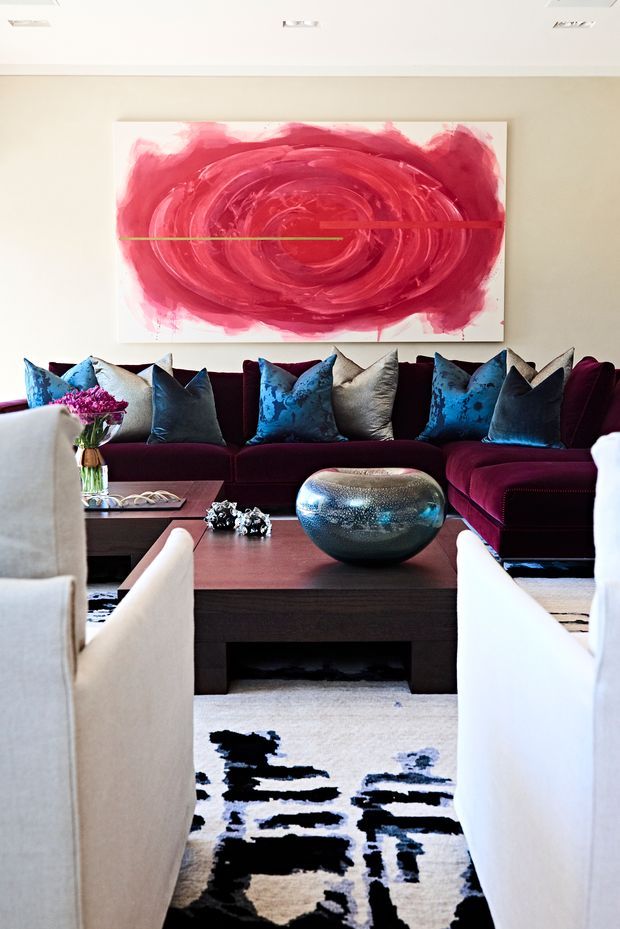 A richly styled living room centred on a deep burgundy sectional sofa with vivid blue, grey, and patterned cushions. Two light upholstered armchairs frame a dark timber rectangular coffee table, topped with a rounded metallic decorative vessel and small accessories. A large abstract red and pink artwork dominates the pale wall, while a bold black, white, and grey patterned rug grounds the seating area. Recessed ceiling lighting and a refined, high-contrast palette create a luxurious contemporary atmosphere.