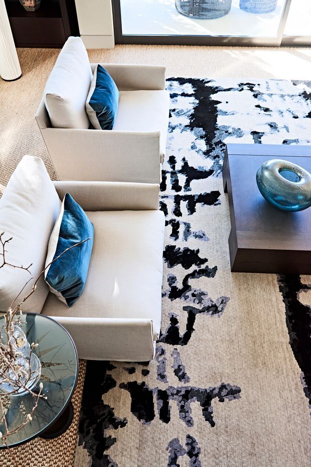 Bright contemporary living room viewed from above, featuring two cream upholstered armchairs with vivid blue cushions, a large abstract black, white and pale blue rug, a dark wood coffee table with a blue glass vessel, and a round glass side table. Floor-to-ceiling glazing brings in abundant natural light.