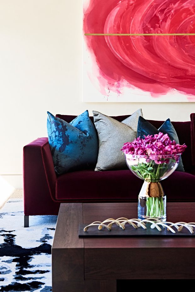 A richly styled living room vignette featuring a deep burgundy upholstered sofa with blue, teal, and grey cushions, a dark timber coffee table, a glass vase of pink flowers, and a large abstract red and pink artwork. A patterned black-and-white rug and strong jewel-tone palette create a dramatic, refined atmosphere.