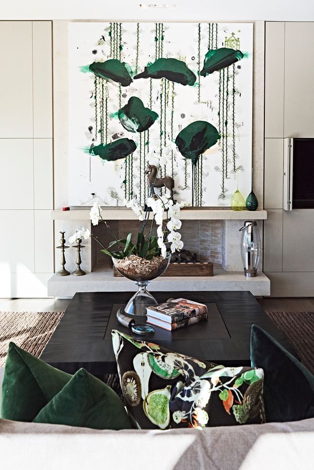 A refined living room centered on a large abstract artwork above a stone fireplace. The room features pale built-in wall cabinetry, a textured stone or tile fireplace surround, a dark rectangular coffee table, a large orchid arrangement, sculptural candleholders, and deep green and patterned cushions on a sofa. The palette combines white, grey, charcoal, green, and natural wood tones with a polished, curated feel.