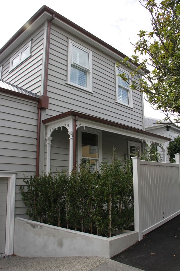 Exterior view of a narrow two-storey weatherboard townhouse with a covered front veranda, decorative fretwork and turned-style porch detailing, white-trimmed windows with shutters, a pale rendered planter wall, and a white vertical-slat privacy gate. The home has a pitched roof with metal roofing and a restrained grey, white and muted red-brown colour scheme.
