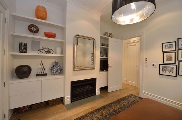 A refined traditional living room or hallway sitting area with built-in white display shelving and cabinetry, a black fireplace, timber flooring, ornate patterned rug, framed artwork, and a large contemporary pendant light. Decorative objects and a mirror add layered visual interest, while a doorway leads to an adjacent hall or room.