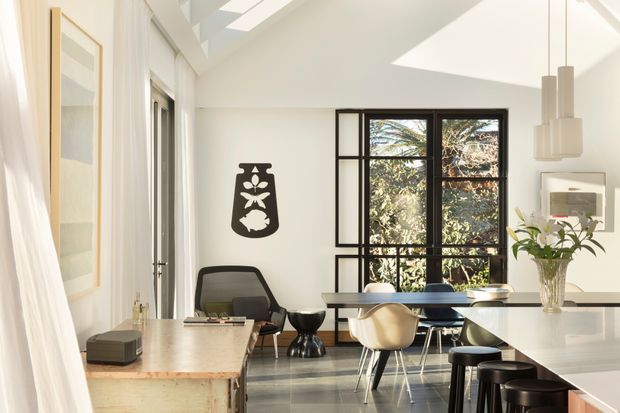 Bright, open-plan contemporary interior combining a kitchen, dining area and living/work zone. The room has white walls and ceiling, large black-framed glazing, a skylight, polished grey flooring, pale timber cabinetry or a timber work surface, and a white island with dark stools. Sculptural pendant lights, modern furniture and indoor plants add visual interest. The space feels airy, refined and connected to leafy outdoor views.