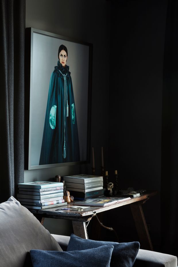 Moody, dark-toned interior vignette featuring a large framed portrait above a rustic console table. The table is styled with stacked books, magazines, candles and small decorative objects, while the foreground includes a light upholstered armchair or sofa with dark blue cushions. Heavy dark curtains and charcoal walls create an intimate, dramatic atmosphere.