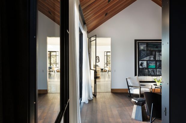 A refined interior view showing a compact living or sitting area opening toward a brighter adjoining space. The room has dark timber flooring, white walls, a warm slatted timber ceiling with recessed spotlights, large black-framed glazing or doors with sheer curtains, and contemporary seating beside a small table. A framed artwork hangs on the right wall, while reflections and the open glazed partition extend the sense of depth.