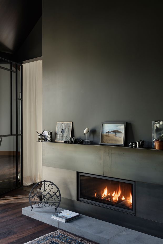 A moody contemporary living room centred on a built-in linear fireplace beneath a long dark grey built-in media or display cabinet. The room features dark timber flooring, a charcoal painted wall and ceiling, black-framed glazing with sheer curtains, a concrete hearth, and a decorative metal lounge chair. Small artworks and objects are arranged along the joinery, with a patterned rug partially visible in the foreground.