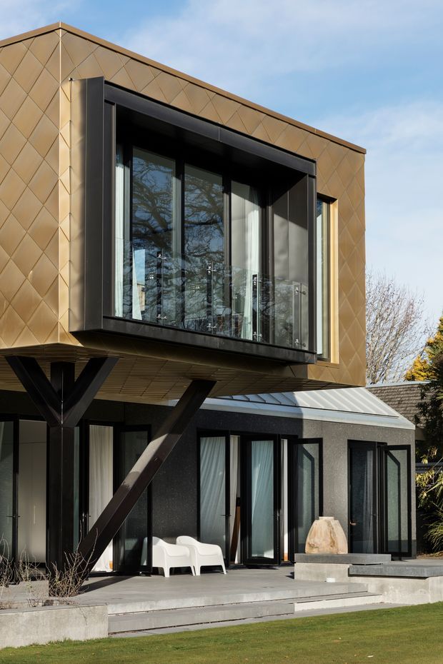Exterior architectural photograph of a contemporary two-storey house. The upper level projects outward over the lower facade and is supported by dark angled structural braces. The facade combines bronze-gold diamond-patterned metal cladding with black-framed floor-to-ceiling glazing and a broad recessed balcony or glazed opening. The ground floor has dark textured walls, extensive black-framed doors and windows, a pale low-pitched roof section, concrete terrace steps, and a small outdoor seating area. The image is captured from the garden, with trees and lawn in the surrounding setting.