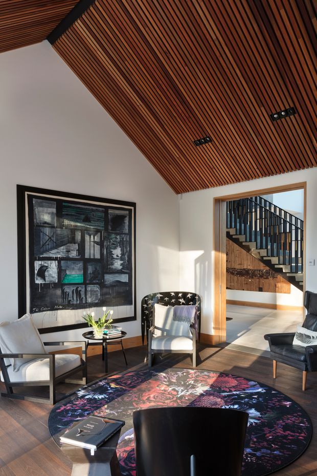 A refined living room with a dramatic pitched ceiling clad in narrow warm timber battens, creating a strong linear feature. The room has pale walls, dark timber flooring, mid-century-inspired lounge chairs, a black-framed abstract artwork, a small side table, and a large round multicoloured floral rug. A wide timber-trimmed opening reveals a modern stairway with black vertical balustrades and timber detailing beyond.