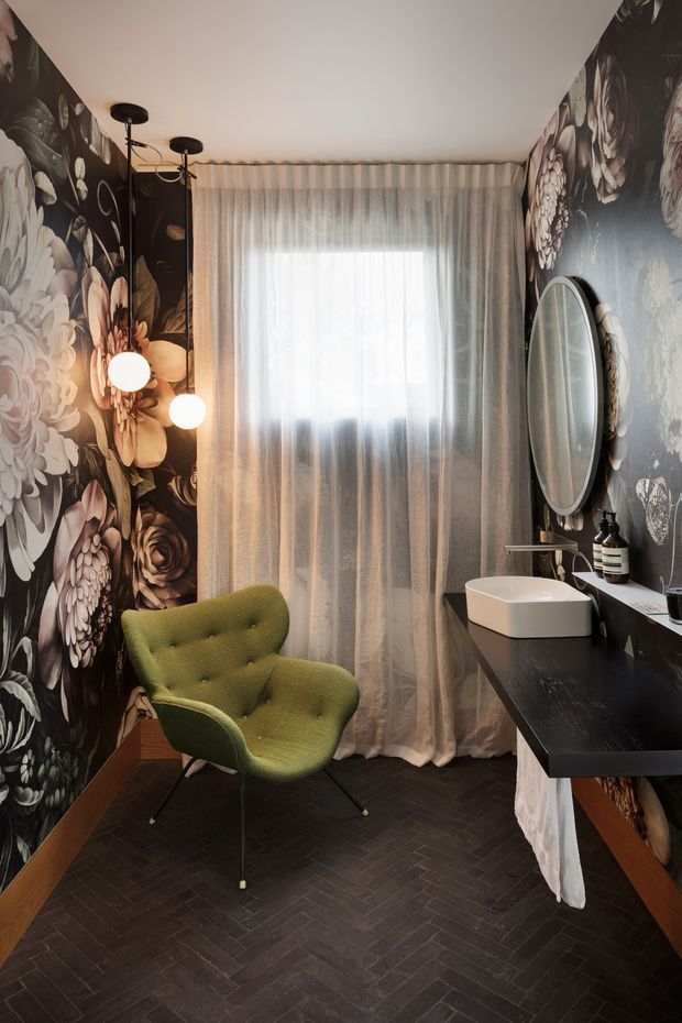 A dramatic, richly styled bathroom with dark floral wallpaper on both side walls, a black floating vanity with a white vessel sink, oval mirror, wall-mounted tapware, and concealed storage. A sheer-curtained window provides diffused natural light, while sculptural pendant lights illuminate a green upholstered accent chair. The floor is finished in a dark herringbone pattern, creating a moody and luxurious atmosphere.