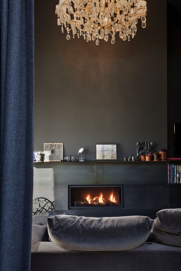 A moody, sophisticated living room focused on a dark fireplace wall with a built-in linear fire, floating display shelf, and deep charcoal cabinetry or panelling. A large ornate crystal chandelier hangs overhead, while a plush dark grey sofa fills the foreground. Blue-grey curtains frame the left side, with books, ceramics, framed art, and copper-toned objects arranged on the shelf.