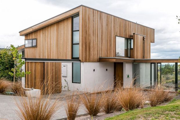 Contemporary two-storey residence with a strong geometric form, extensive vertical timber cladding, white rendered lower walls, dark-framed glazing, and a low-pitched roof. The house is set within landscaped ornamental grasses, with a covered outdoor terrace extending from the ground floor.